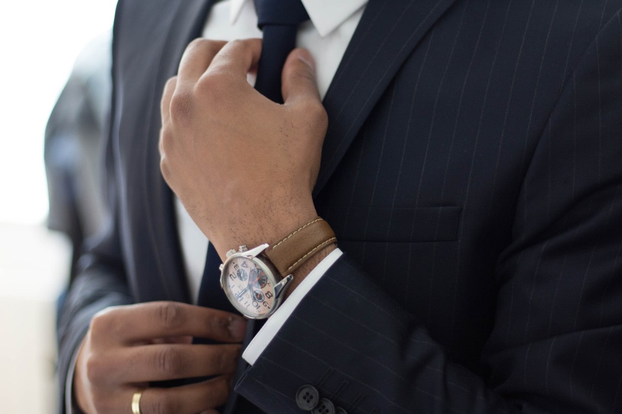 A man in a suit adjusting his tie, showing off his luxury watch with a light brown leather band.