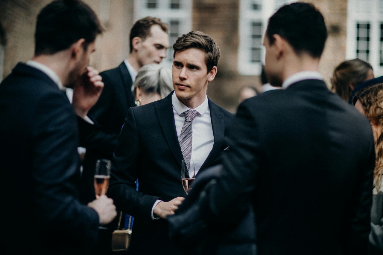 A few men in suits drinking champagne and having a discussion at an outside group gathering.