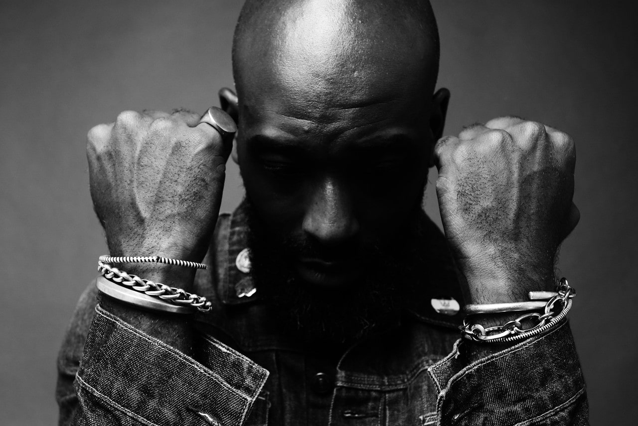 A black and white photo of a man wearing a denim jacket with his hands raised in fists, displaying his many bracelets of different textures and chain sizes.