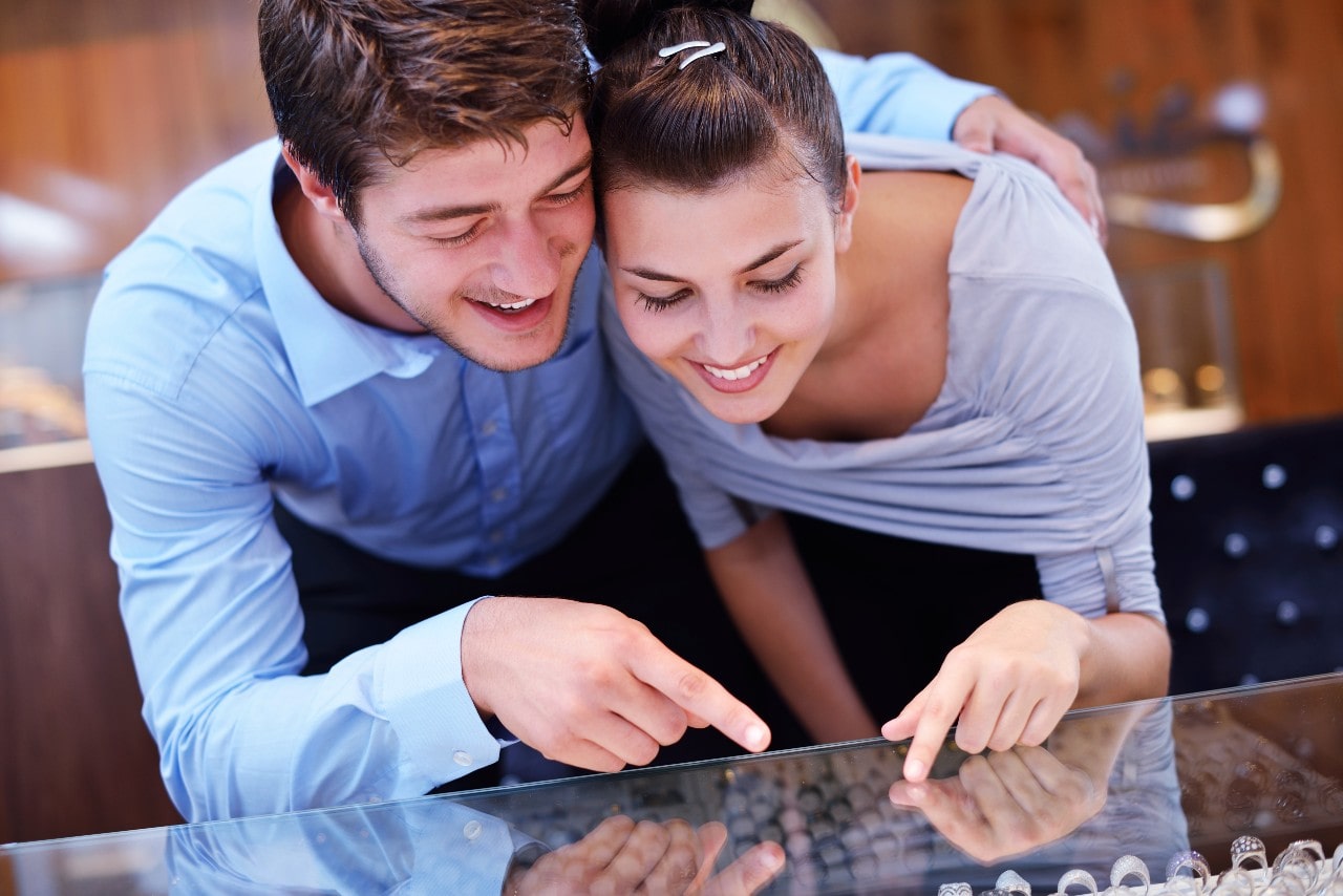 A smiling couple leans over a jewelry display case, excitedly pointing at rings inside.