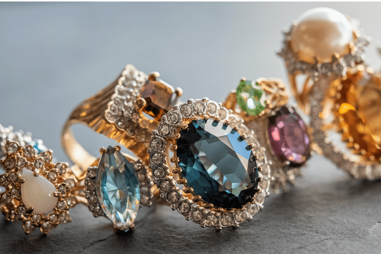 A close-up of ornate gold rings featuring large faceted gemstones in blue, green, pink, and amber tones, each surrounded by sparkling diamond accents.