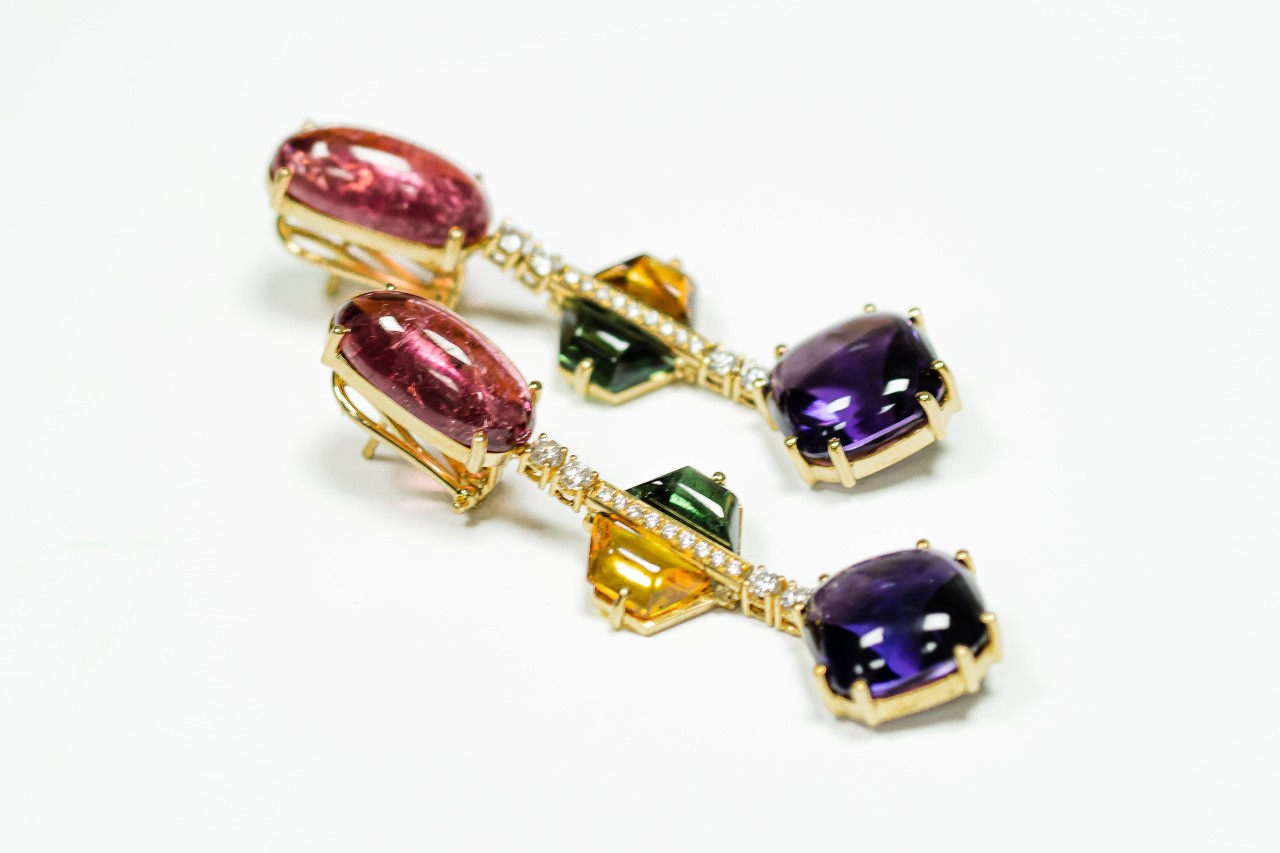 A close up of dangle gemstone and diamond earrings in yellow gold, featuring rubies, sapphires, yellow and green crystals.