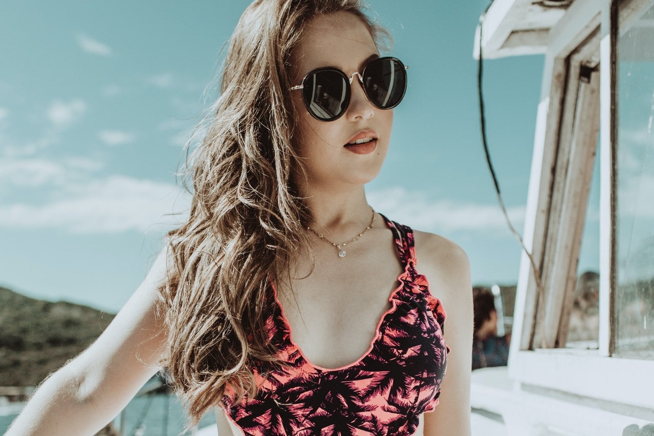 A close-up of a woman on a boat wearing round black sunglasses, a yellow gold chain necklace, and a pink patterned swimsuit.