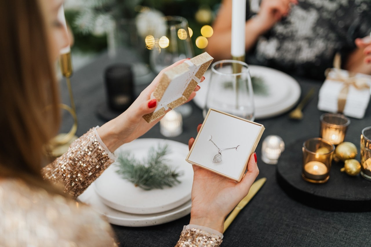 A woman in a sequined dress opens a gift box containing a sparkling diamond pendant necklace at a festive dinner table.