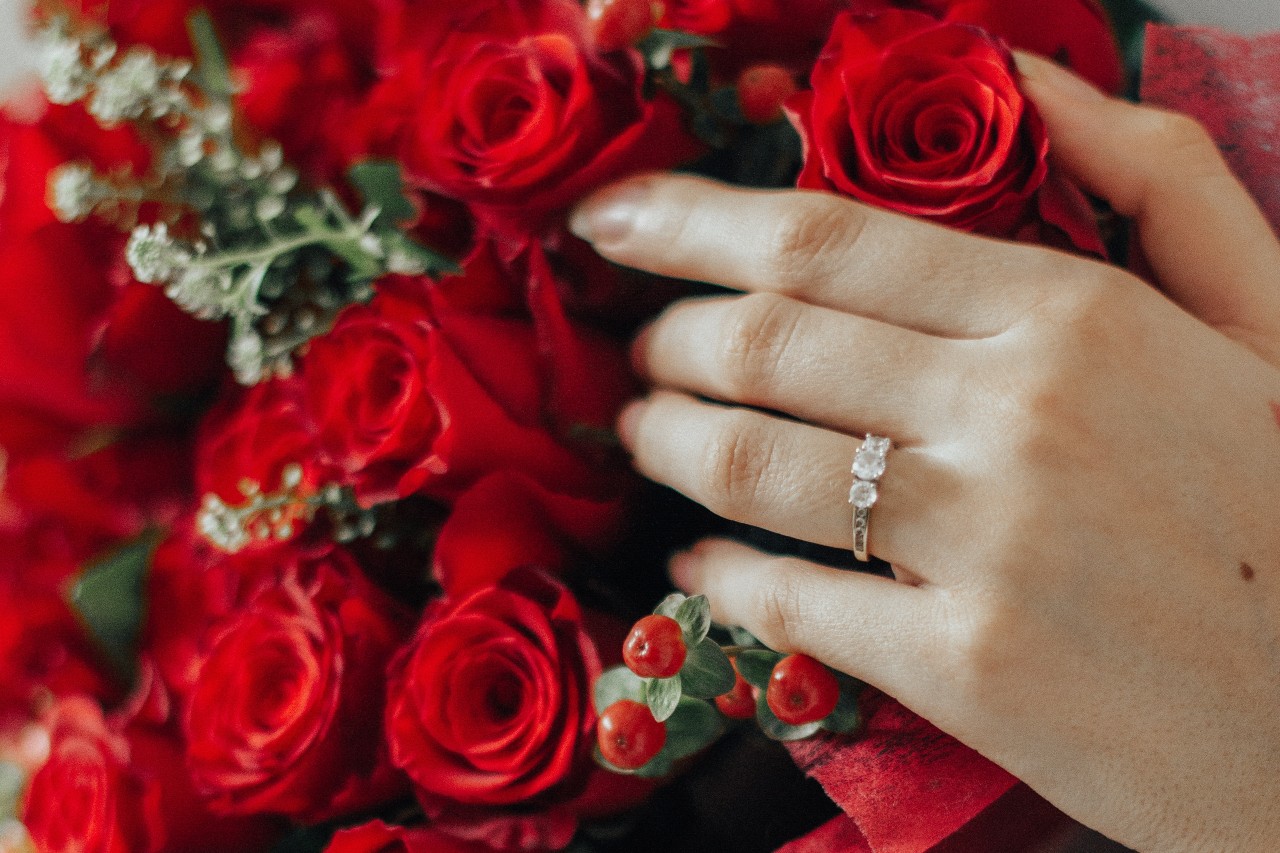 A three stone diamond ring on a woman&rsquo;s hand resting on a bouquet of red roses