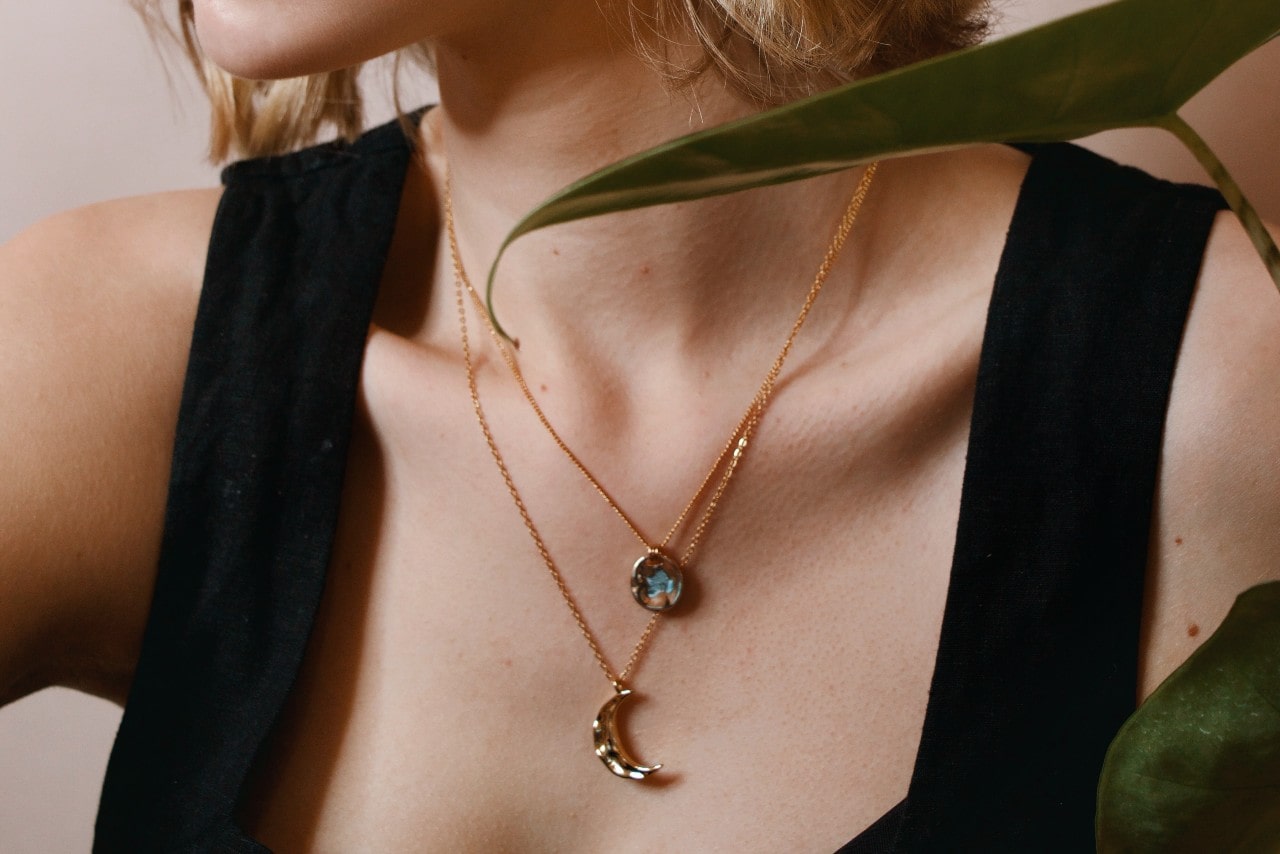 A woman in a black dress wears layered gold necklaces with moon and gemstone pendants.