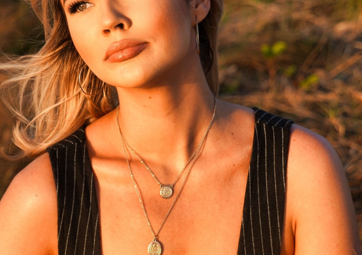 Woman in a striped top, with gold necklaces and hoop earrings, gazing upward in warm sunset light.