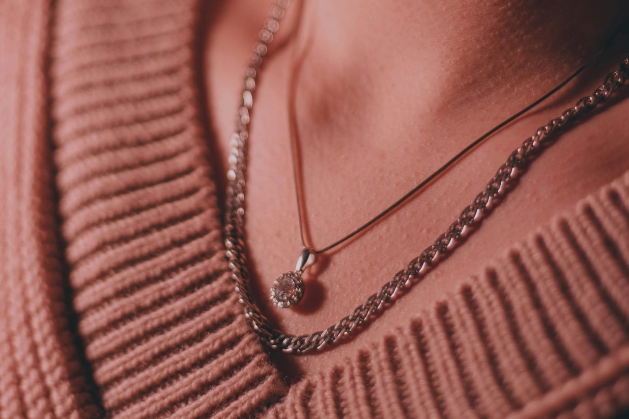 Close-up of a woman wearing a silver chain necklace and a pendant on a thin cord, both against a ribbed, pink sweater.