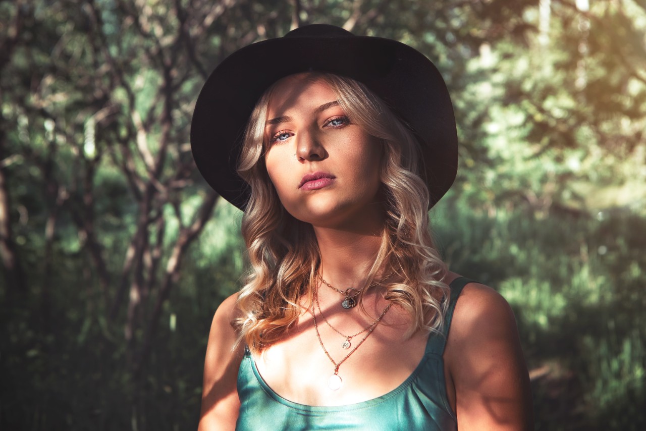A woman with curly blond hair stands in front of the forestry backdrop, showcasing layered necklaces.