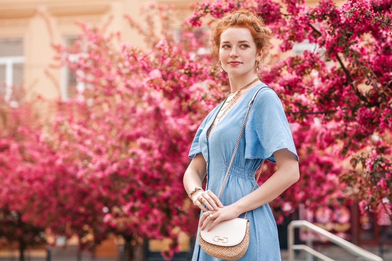 A woman in a blue dress and with ginger hair stands in front of the blooming pink trees.
