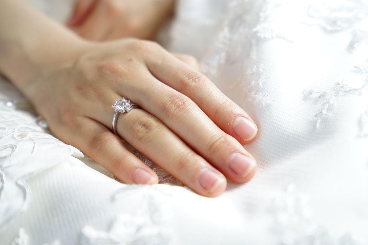 A white gold diamond ring on a woman&rsquo;s hand on top of the white embroidered fabric.