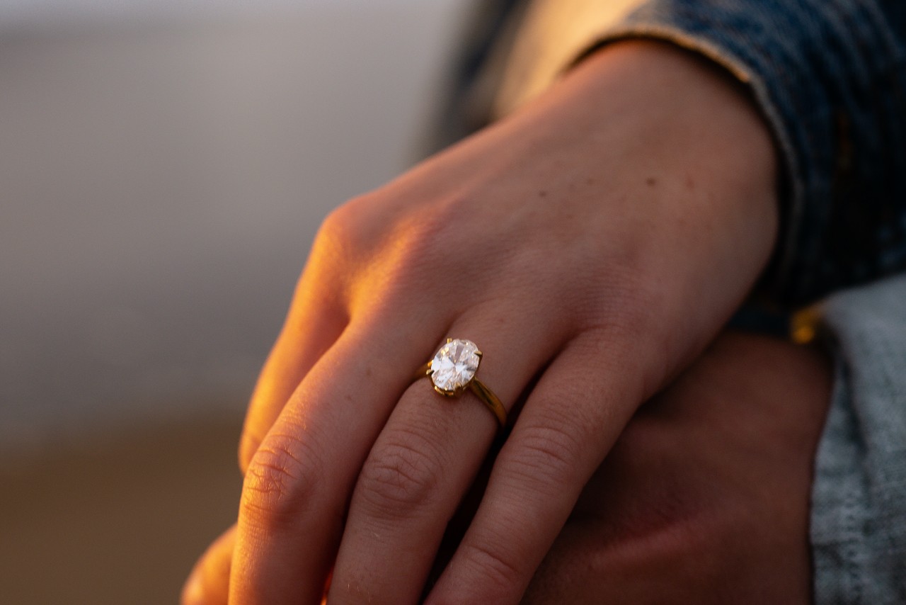 a woman’s hand lying over another’s hand, and wearing a yellow gold solitaire ring with an oval cut diamond