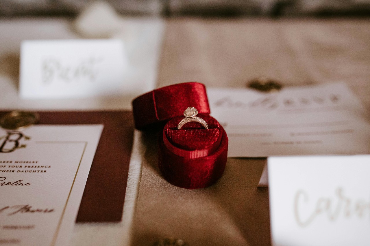 a diamond ring in a red ring box on a table with wedding invites and place cards.