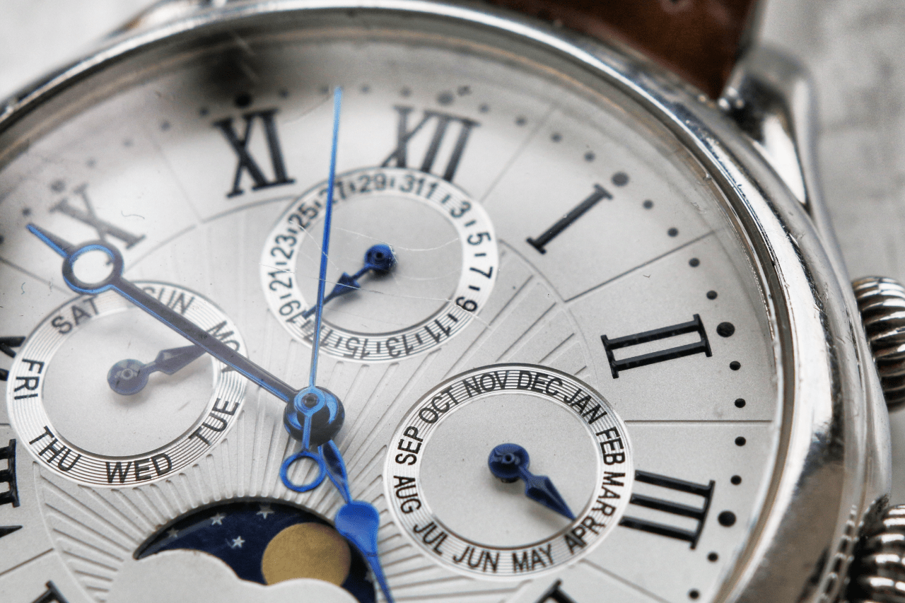 A close-up of a watch face featuring Roman numerals and multiple complications, with blue hands and detailing.