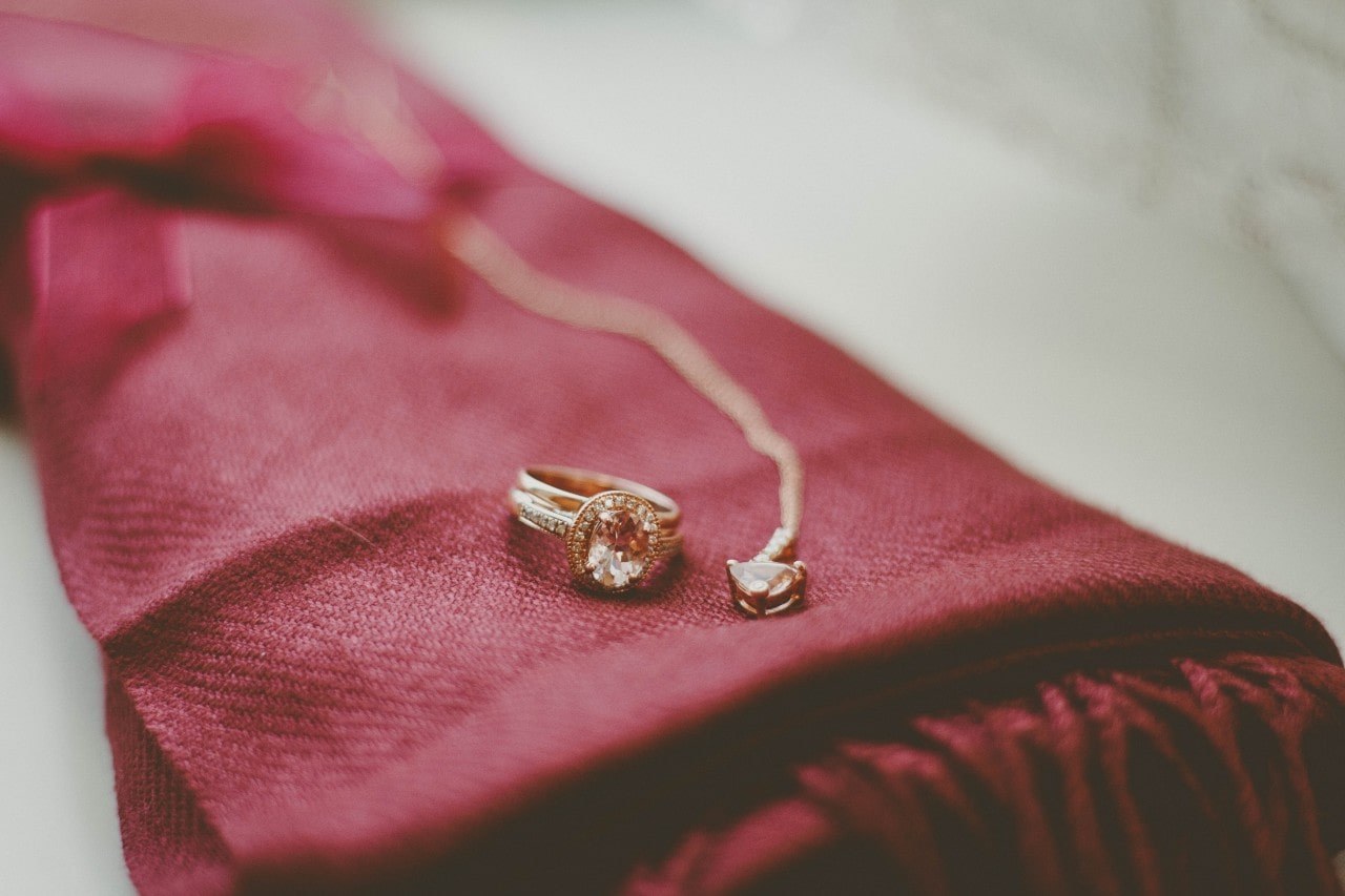 A oval cut halo ring sits on a burgundy fabric beside a gold pendant.