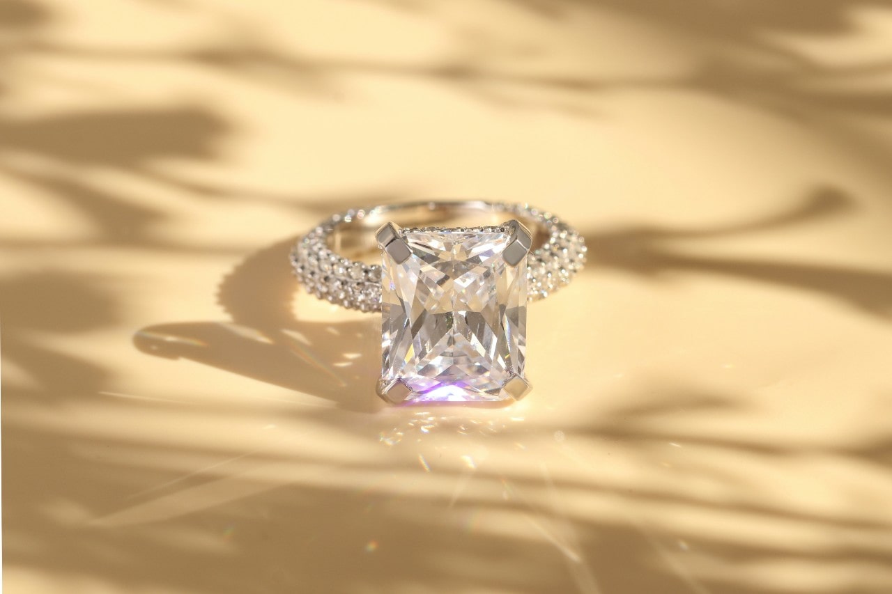 A radiant cut diamond engagement ring sits on a tan table outside.