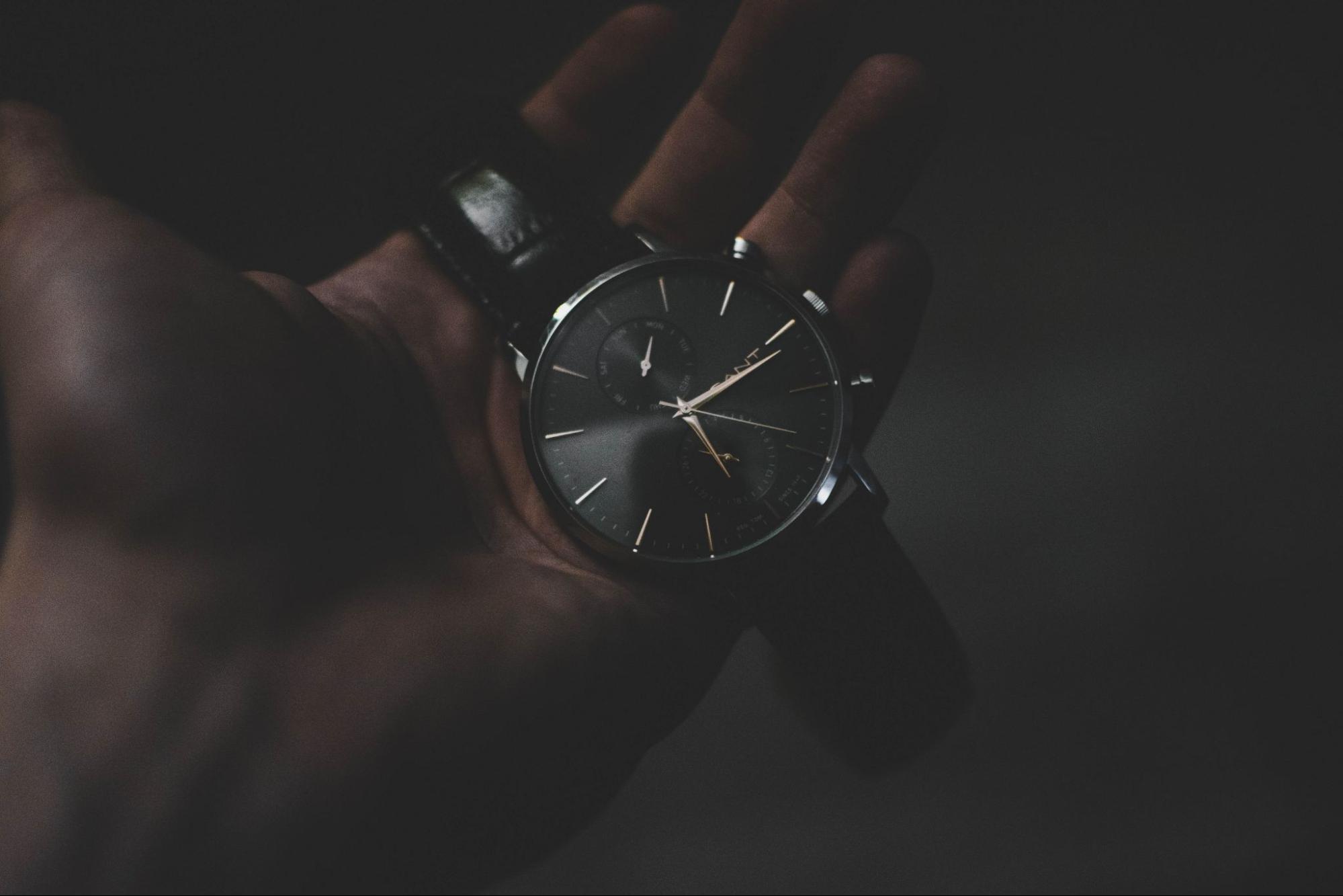 A man holds his black minimalist watch in a dimly lit room.