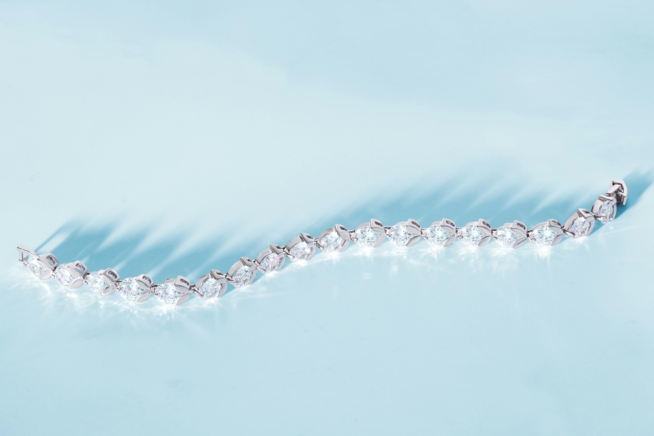 Silver bracelet with sparkling gemstones arranged in a wavy line on a light blue background, casting soft shadows.