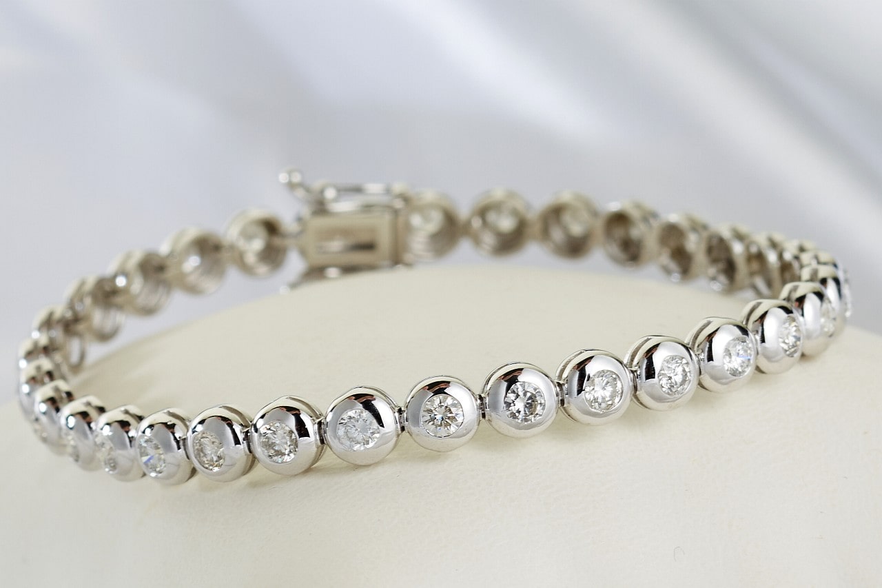 A silver bracelet with round, sparkling diamonds set in bezel settings, displayed on a soft, white surface.