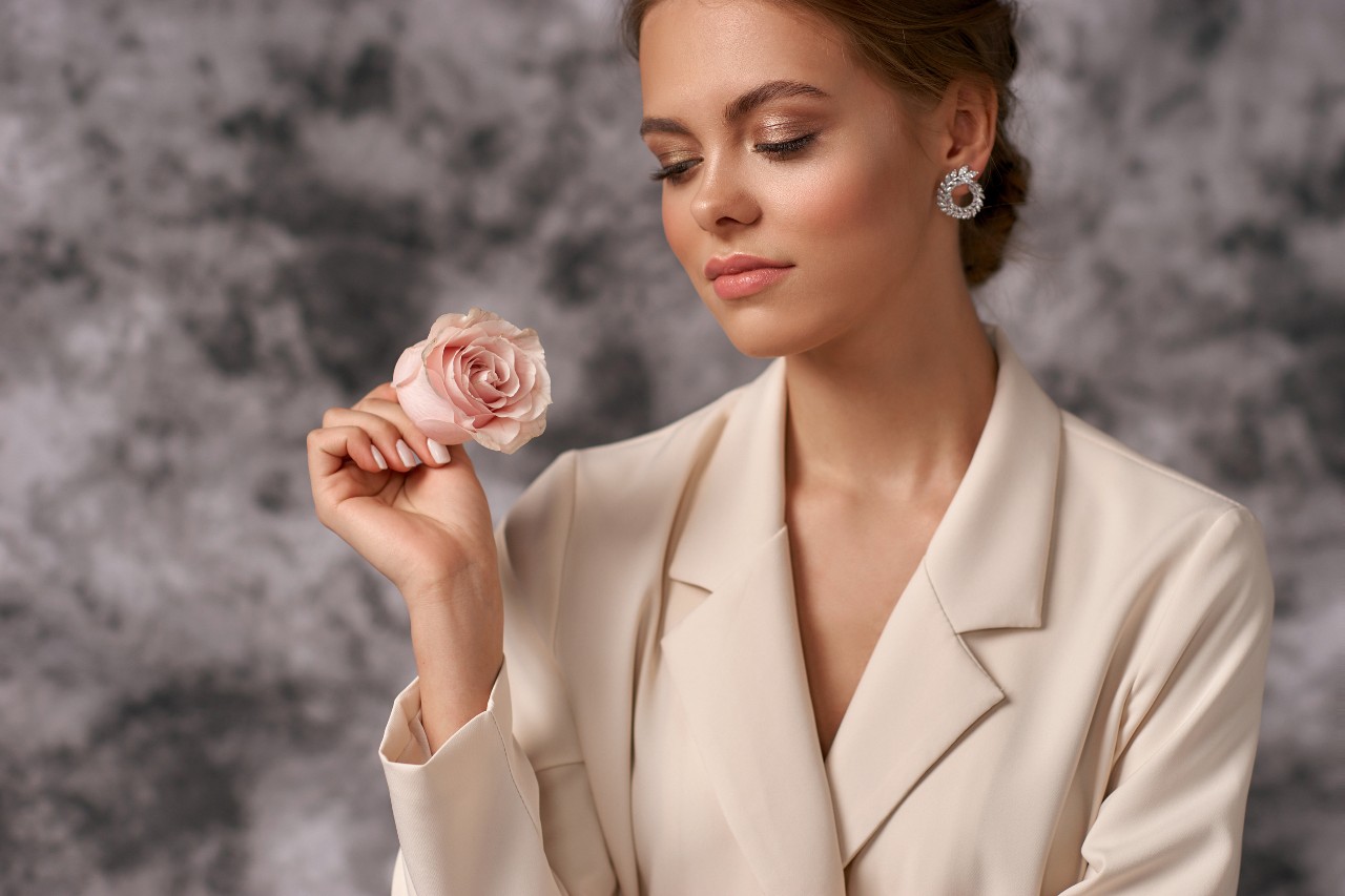 A woman in a cream blazer and diamond earrings holds a pink rose, posing gracefully against a blurred gray background.