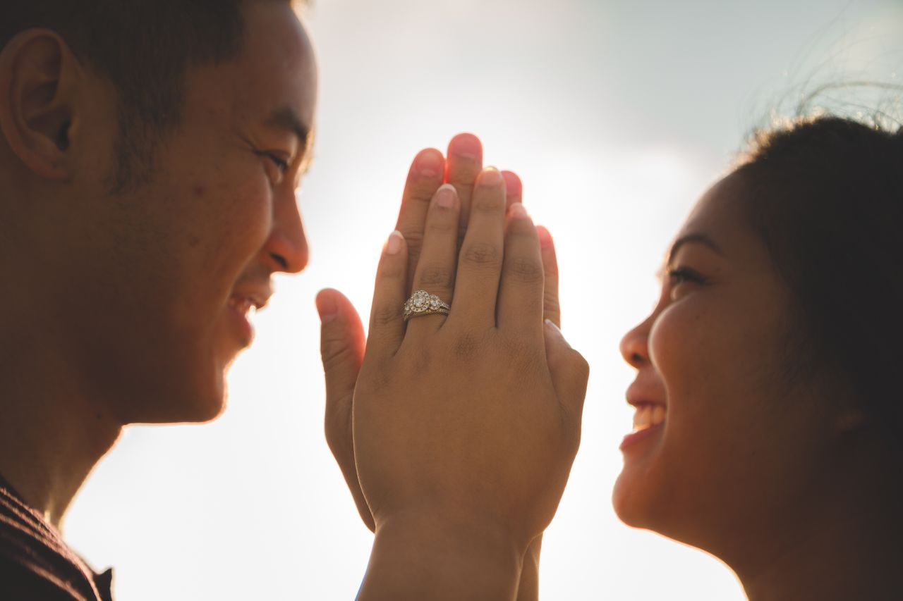 a couple smiling at each other and holding up their hands, the woman wearing a three stone halo engagement ring