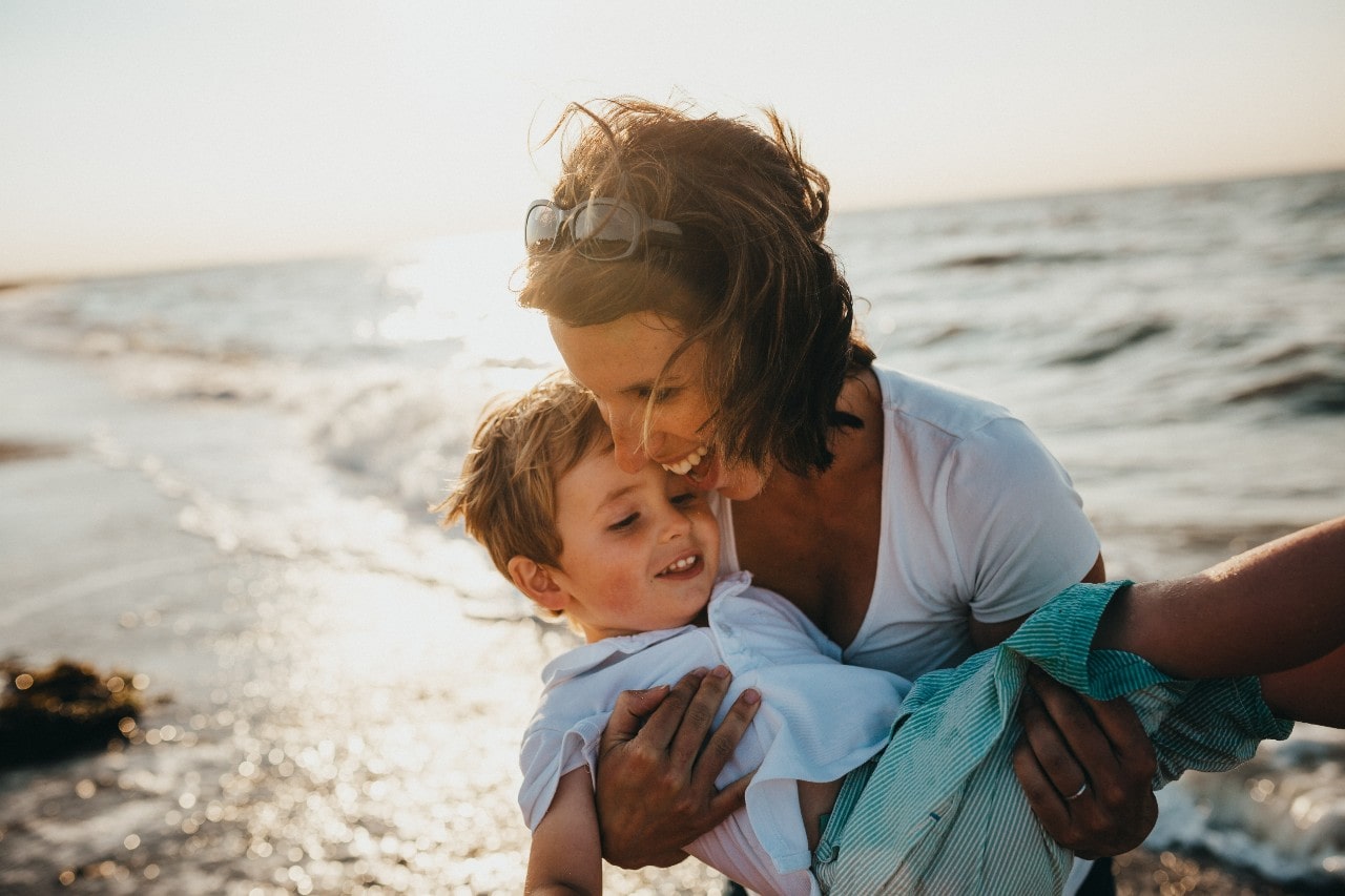 A mother holding her son joyously on the beach, the sun shining in the background.