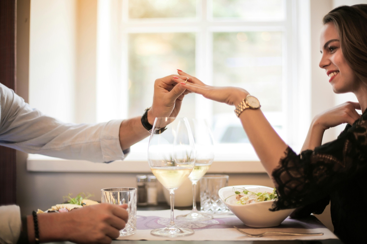 A man in a white shirt and black leather wristwatch putting an engagement ring on a woman’s hand, who is wearing a yellow gold metalling wristwatch and a black lace blouse, while dining at the restaurant.