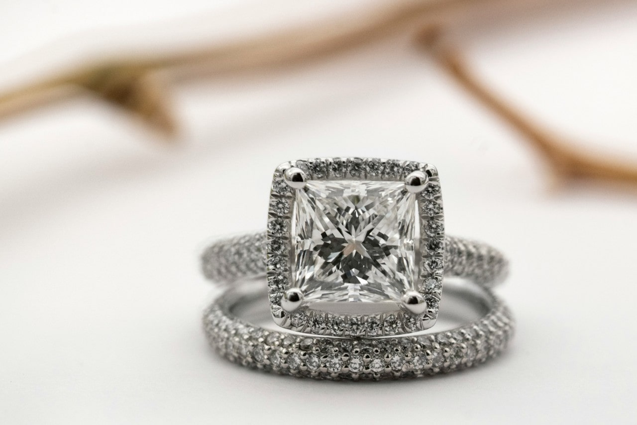 A close-up of a dazzling cushion-cut diamond engagement ring with a matching diamond-studded wedding band, displayed on a white surface.