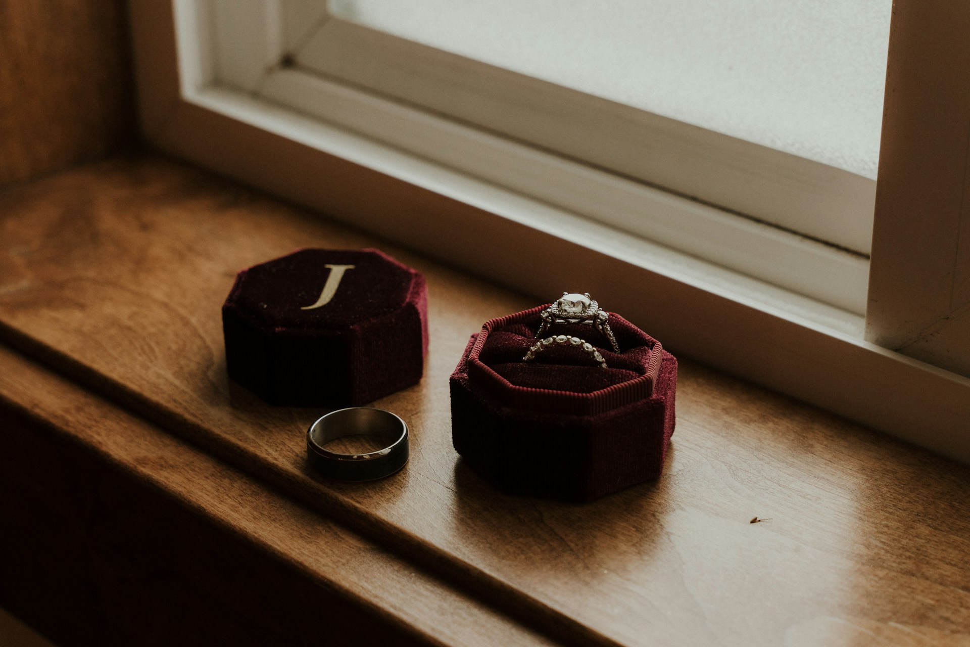 A close-up of a bridal ring set in a red velvet jewelry box placed on a wooden surface near the window.