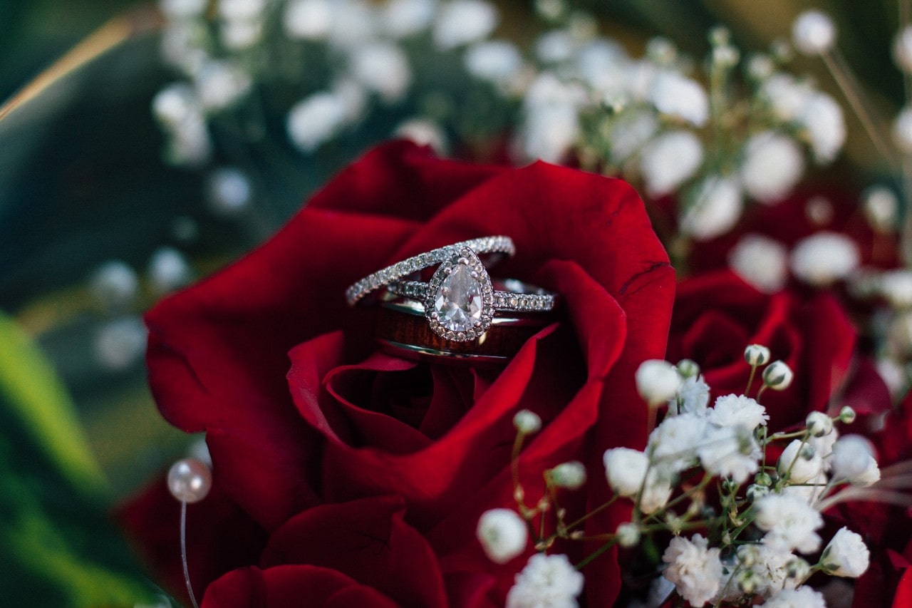 A teardrop diamond ring with a sparkling band is elegantly placed on a lush red rose surrounded by delicate white baby's breath flowers.