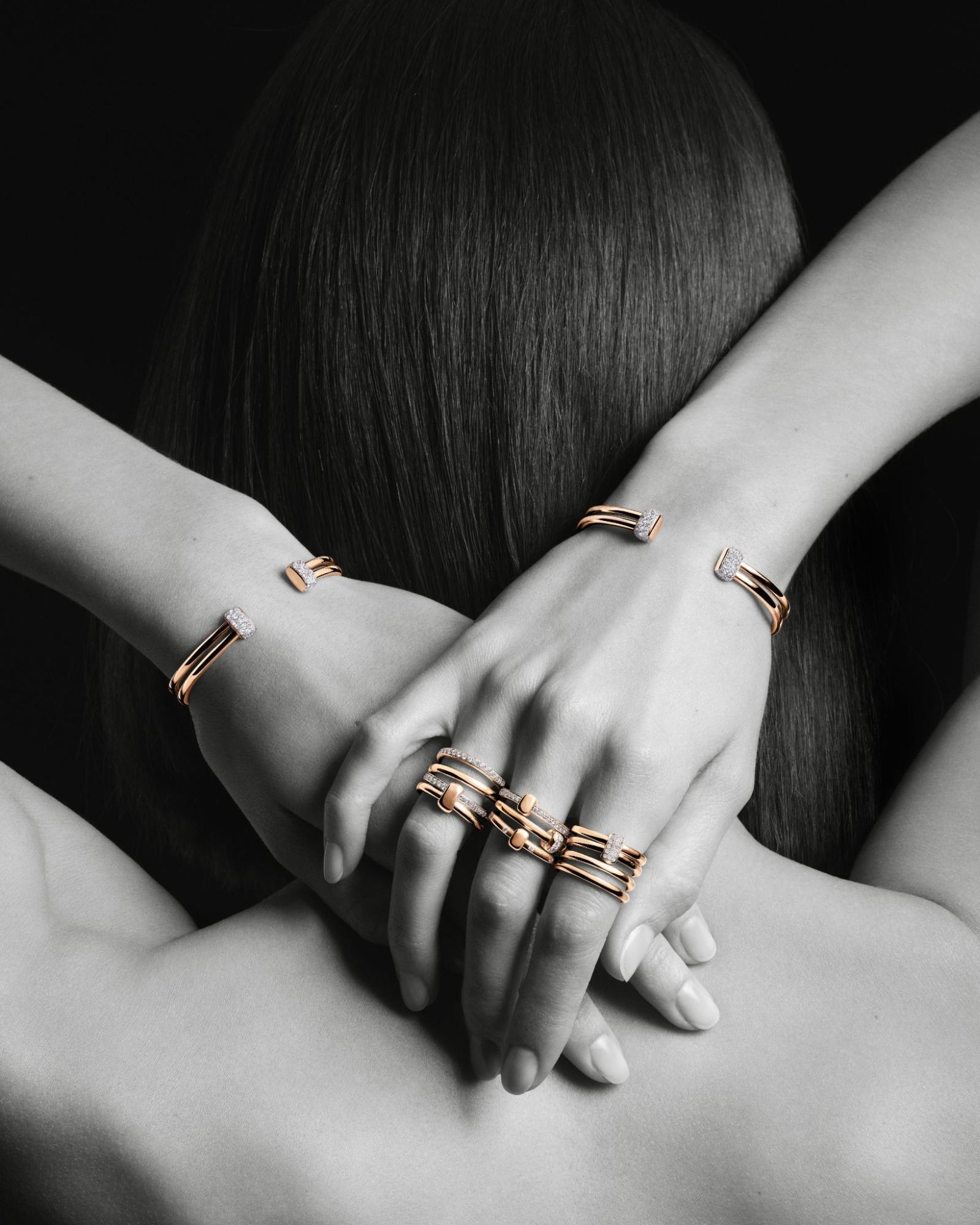 A black and white photo of a woman with yellow gold and diamond rings on her hand and bracelets on her wrists as she holds her arms over her head and hands behind her neck.