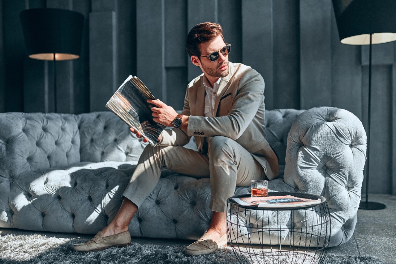 Man in a beige suit and sunglasses sits on a tufted sofa, casually holding a magazine. A luxury watch peeks from one wrist, while a drink rests on a wire table completes the laid-back, upscale scene. Man in a beige suit and sunglasses sits on a tufted sofa, casually holding a magazine. A luxury watch peeks from one wrist, while a drink rests on a wire table completes the laid-back, upscale scene.