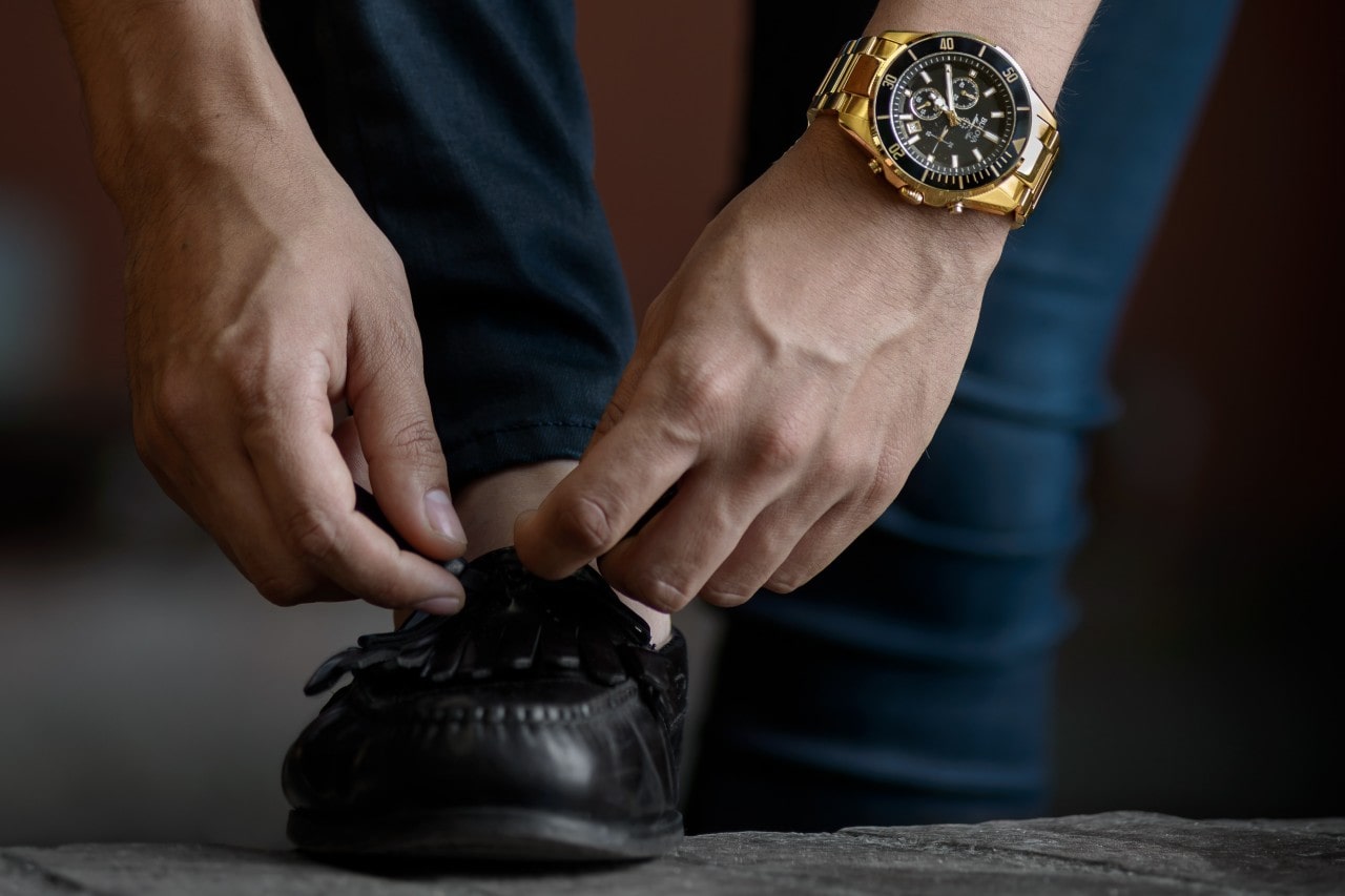 A person wearing a gold watch ties the laces on a black leather shoe. The focus is on the hands and watch, conveying a sense of detail and style. A person wearing a gold watch ties the laces on a black leather shoe. The focus is on the hands and watch, conveying a sense of detail and style.