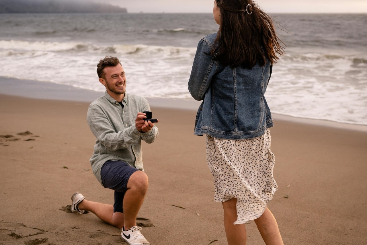 A man is proposing on a beach, kneeling with a ring box open while smiling at a surprised woman standing in front of him.
