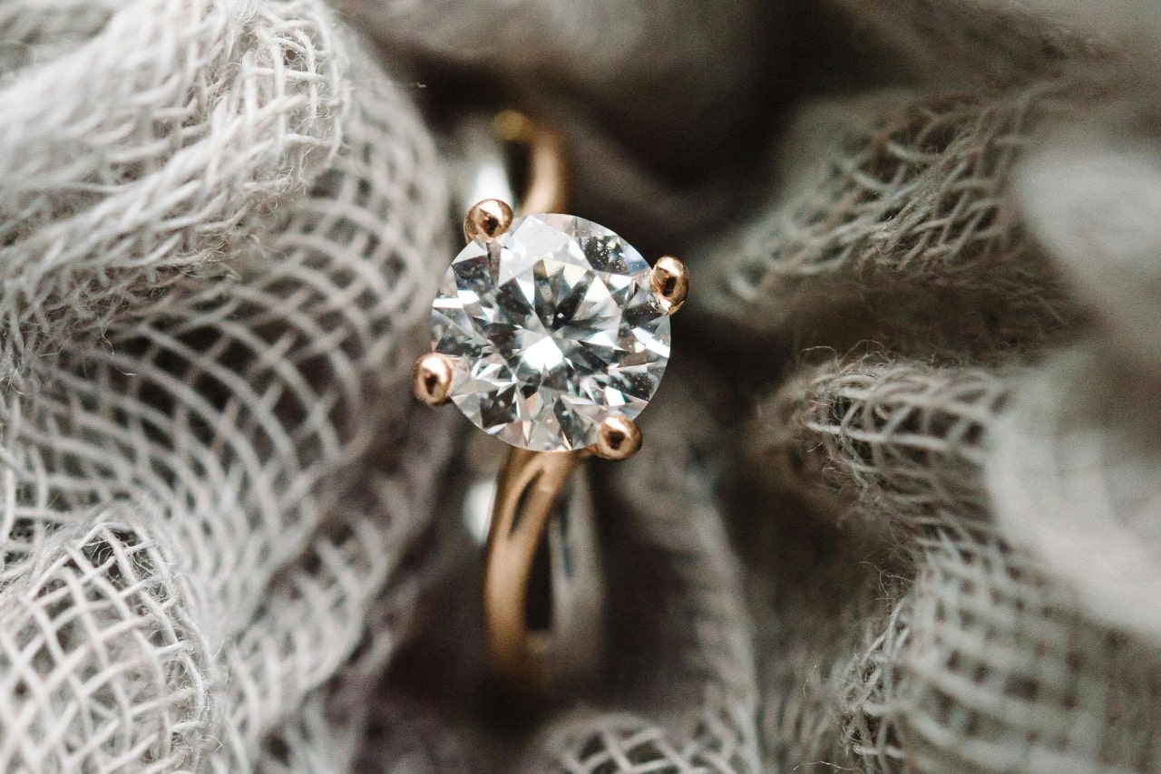 A close-up shot of a gold engagement ring with a round-cut diamond in soft, textured fabric.